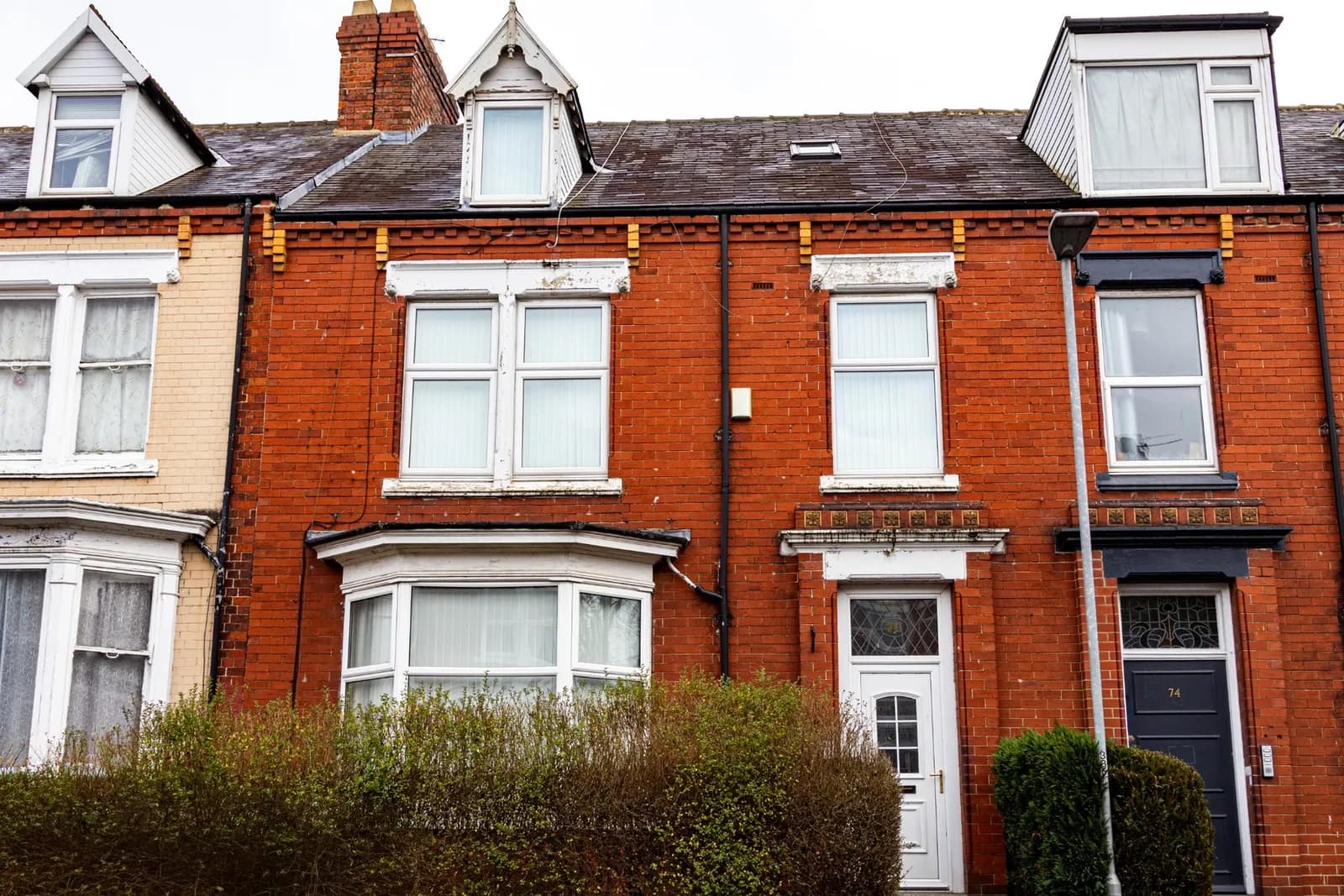 A row of terraced brick houses with various windows and dormers, and a hedge in the foreground.