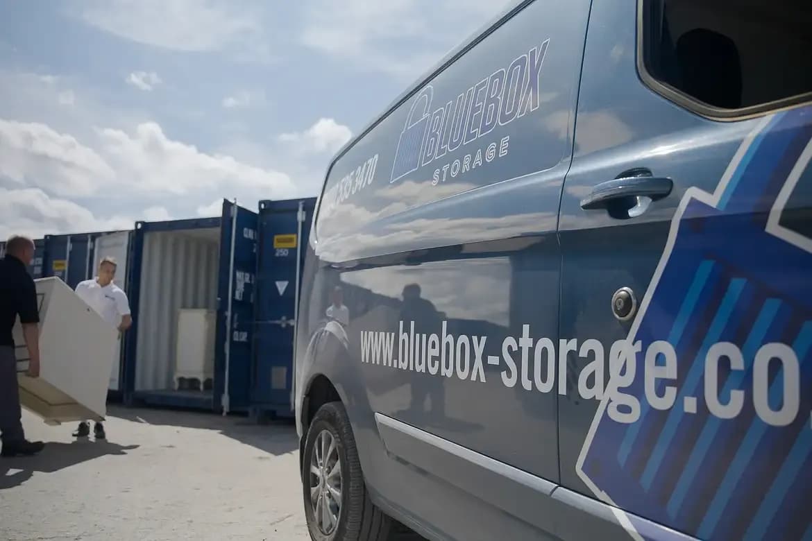A close-up of a blue Bluebox Storage van with its website address on the side, and in the background, two men are loading a large white item into a blue shipping container.