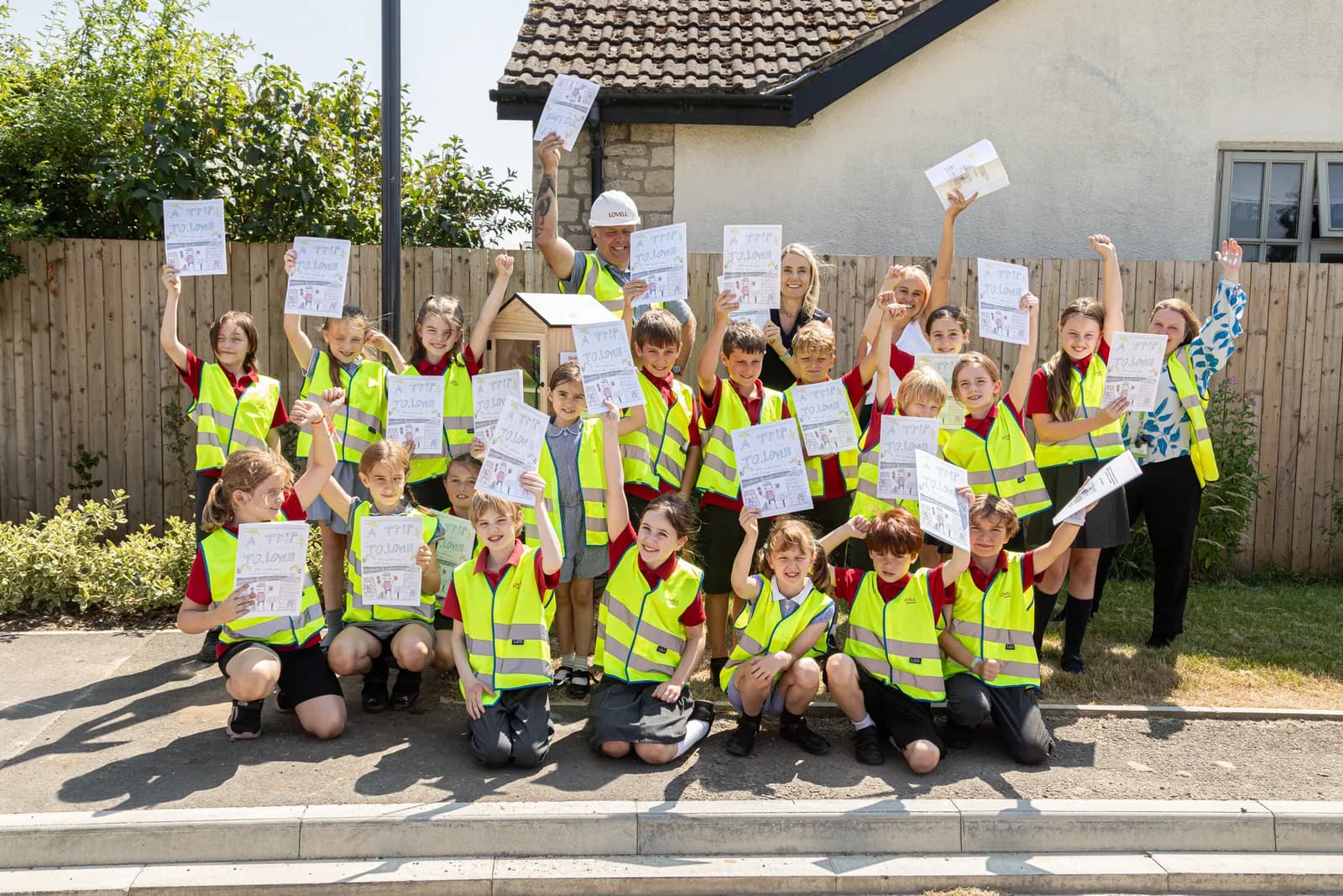 A group of children and adults, all wearing high-visibility vests, stand and kneel outdoors, holding up drawings and smiling at the camera.